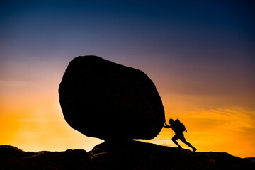 Silhouette of man with backpack over blue sky and orange background pushing huge rock