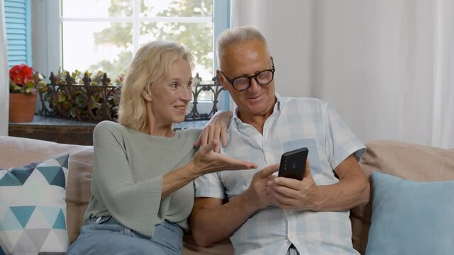 Happy elderly couple relax on couch using cellphone