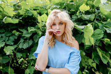 Young beautiful blonde girl in a blue dress bites her finger and touches her lips with her hands. Naughty, funny and playful woman in the park.