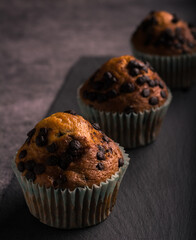 
Three delicious muffins with pieces of chocolate on a black slate. Gray background. Vertical format