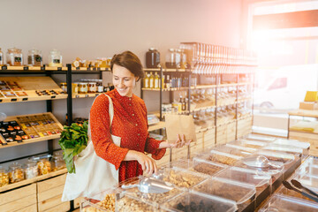 Young woman in red dress with reusable cotton bag doing shopping in plastic free store. Minimalist vegan girl buying nuts, groceries without plastic packaging in zero waste shop. Low waste lifestyle.