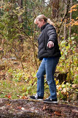 Fototapeta premium This midlife aged Caucasian woman is balancing while walking on a log in the forest on a cold, damp day. She's wearing blue jeans, and a black down coat. 