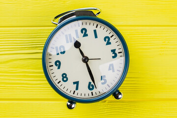 Close up round clock with reflective glass. Clock hand shows half past eleven. Yellow wooden background.