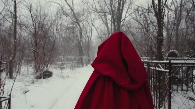 Little Red Riding Hood. A girl in a red coat with a hood walks through a snowy cemetery in winter. Snowing in slow motion