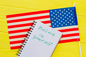 Believe in yourself phrase written on the notepad. Close up us flag on yellow wooden table.
