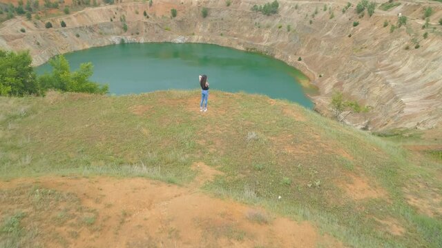Young woman standing on cliff edge and looking at abandoned copper mine pit partially full of blue water