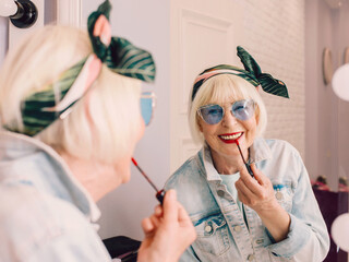 Smiling elderly senior stylish woman in blue sunglasses and jeans jacket using red lipstick by the mirror in stylish loft interior. Style, fashion, make up, anti age concept