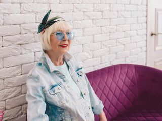 elderly senior stylish woman in blue sunglasses and jeans jacket sitting on the sofa in stylish loft interior in salon. Style, fashion, wait, anti age concept