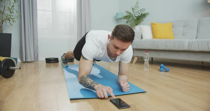 Muscular guy doing fitness and working out on a mat, listening to music in the wireless earphones. Athlete with lots of stamina doing a plank exercise using a smartphone app, timing in a stopwatch.