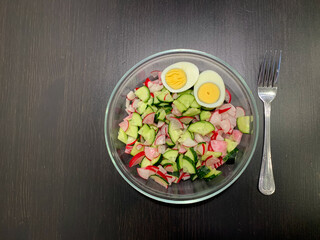 In the photo, a Salad of chicken egg, radish, cucumber on a dark wooden table in a transparent plate with a fork. Photo from above. Proper nutrition, vegetarian diet.