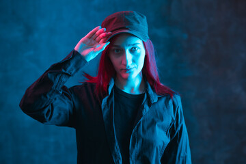 Red haired woman in cap and black shirt on dark background with colorful neon lighting.