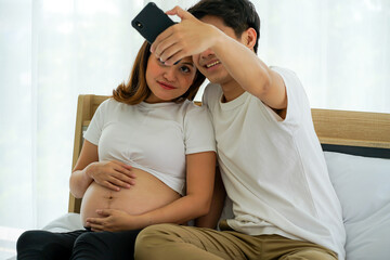 young Asian pregnant woman with husband on the white bed in the house taking selfie  with mobile cellphone
