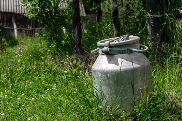 An old metal can (barrel with a lid) for water, stands on the ground, overgrown with green grass.