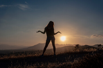 Silhouette of a girl in a black T-shirt, jeans shorts and hats stands with her hands apart on the top of the hill against the backdrop of the sunset and mountains
