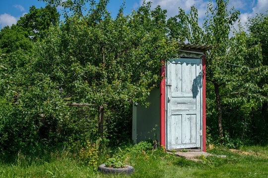 A rustic toilet stands outside among garden trees and green grass.
