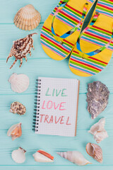 Different sea shells and yellow sandals in the corner on turquoise background. Love live travel written on notepad.
