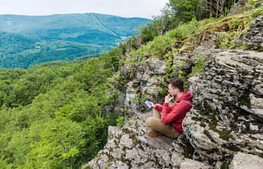 Naklejka premium Young man in a red sweater and beige pants sits on top of a high cliff and reads an e-book. Green forest is spread down below. Low mountains overgrown with tall trees can be seen on the horizon.