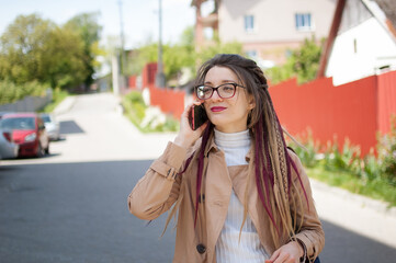 Modern young business woman with long dreadlocks and eyeglasses is talking with the client by mobile phone standing at the street near the buildings