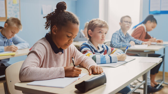 In Elementary School Classroom Brilliant Black Girl Writes in Exercise Notebook, Taking Test and Writing Exam. Junior Classroom with Diverse Group of Children Working Diligently and Learning New Stuff