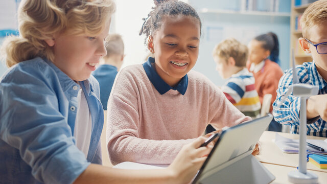 In The Elementary School: Girl And A Boy Work As A Team Using Tablet Computer. Diverse Classroom With Kids Learning Programming Language And Software Design