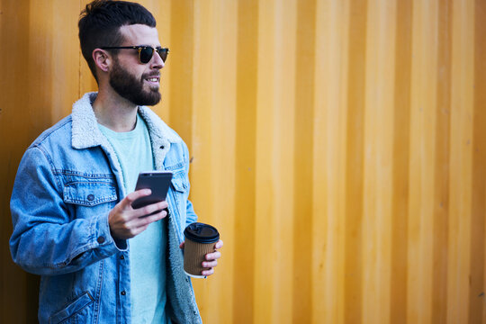 Handsome male hipster in eyewear strolling on city street on vacation trip blogging site motivating followers to travel sharing photos from tour using modern smartphone and fast internet in roaming
