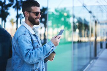 Attractive bearded male entrepreneur dressed in trendy casual look enjoying weekends strolling on city street taking selfie near modern constructed business center making check-in in social networks