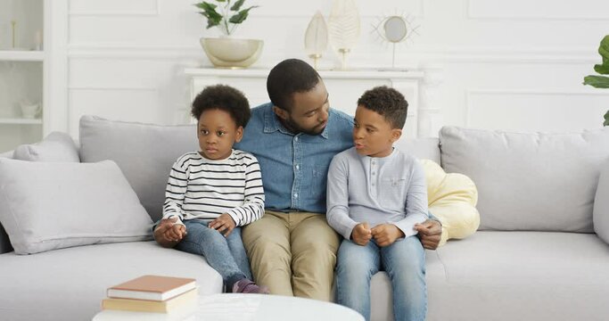 Young African American handsome father sitting on couch with two small kids and talking. Dad hugging little children on sfa and telling them fairytale or preaching. At home. Man with son and daughter.