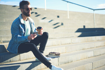 Young handsome international student in casual street wear outfit  enjoying coffee on break in university spending time outdoors chatting with classmates via smartphone and 5G wireless connection