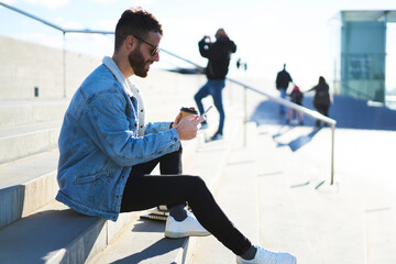 Young male hipster traveler dressed in street wear  enjoying trip during day off taking selfie and posting picture in social networks to share impressions with friends using smartphone and 4G internet