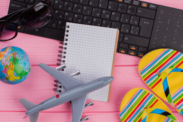 Blank notebook with woman's traveler accessories glasses wallet and flip-flops on pink table top background. Globe black keyboard grey airplane.