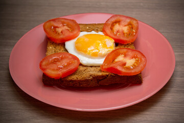 Toast bread with fried egg and sliced tomatoes on pink dish on wooden table.