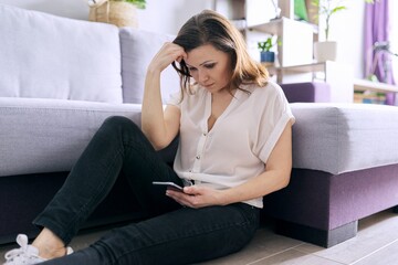 Mature sad woman reading smartphone sitting at home on the floor