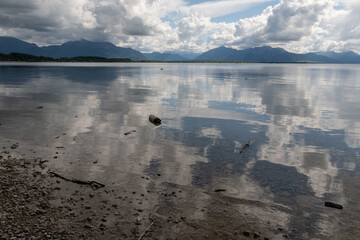 Fototapeta premium Spiegelungen Chiemsee mit Bergen , Himmel und Wolken und schwimmenden Baumstämmen