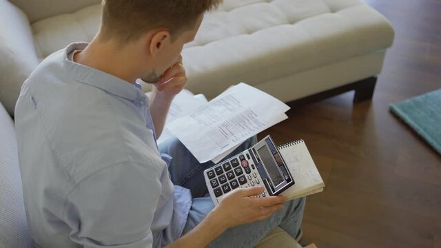 From above shot of perplexed middle aged man sitting on sofa and calculating finances at home. Man counting expenses, thinking with his hand on chin and rubbing forehead