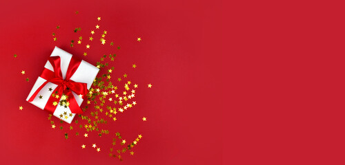 Minimalist Christmas composition with gift box, golden confetti on red background. Greeting concept for New Year, Valentines day, Womens day or birthday.  Top view.  Copyspace.
