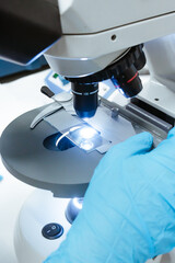 Scientist's hands holding a glass slide in a medical microscope with blue gloves.