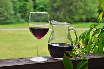 Glasses with red wine and a jug of red wine on a railing terrace in the middle of a forest. Wooden railing overgrown with green leaves.