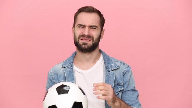 Young sad guy 20s football fan cheer up support favorite team hold soccer ball in denim jacket white t-shirt isolated on pink background studio. People emotions sport family leisure lifestyle concept