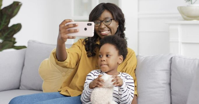 Beautiful young African American mother sitting on couch wih cute little daughter and taking selfie photos with smartphone camera. At home. Woman wih small kid making pictures selfies wih phone.
