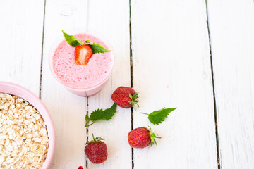 Cocktail with strawberries and milk in a glass decorated with mint and strawberries. Nearby is a pink cup with Hercules flakes, and loose strawberries on a white wooden background.