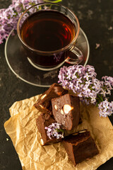 A piece of homemade chocolate cake on crumpled craft paper, with nuts on a black texture table. A cup of tea and lilac flowers decorate a delicious tea party.