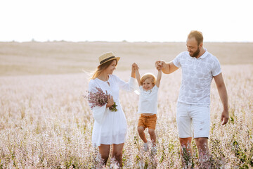 Fototapeta premium Happy family portrait outdoor at the sunset time. People having fun on the field. Concept of friendly family and of summer vacation. Parents and son spending good time. White dress. Straw hat.