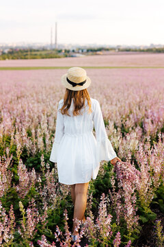 Back view of a pretty young woman in white dress holding straw hat running walking at the sage flower field. Beautiful girl enjoying a field of flowers, relaxing outdoors, having fun, harmony concept.