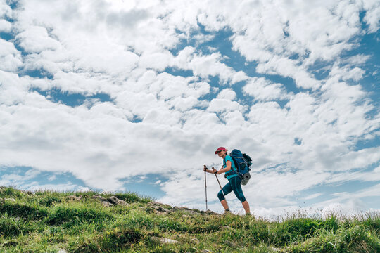 Female backpacker climbing to the mountain top using trekking poles with bright cloudscape background. Active vacation spending by sporty people concept horizontal image.