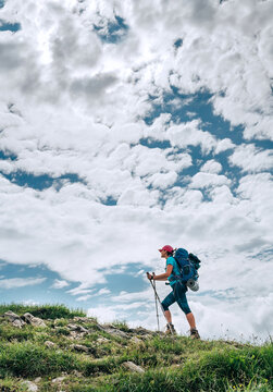 Vertical image of female backpacker climbing to the mountain top using trekking poles with bright cloudscape background. Active vacation spending by sporty people concept image.