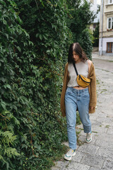 Young woman in casual fashionable outfit walking at green bush wall in city street.Stylish hipster girl walking in european street.  Alone leisure activity in city