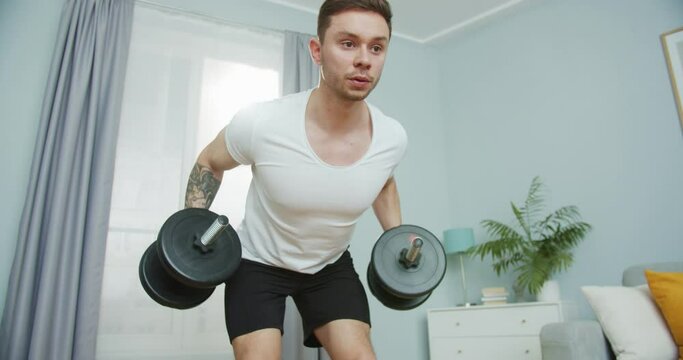 Close up young handsome caucasian sportsman pumping up at home. Strong athletic fit male doing bent over barbell row exercise with dumbbells, weights in the living room. Fitness and sports concept.