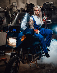 Obraz na płótnie Canvas Young blond woman mechanic in work overalls hold big wrench while sitting on sportbike in garage or workshop, smiling and looking on camera
