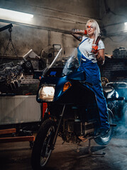 Fototapeta na wymiar Beautiful female mechanic with tattooed hands wearing work overalls posing on her sportbike in garage or workshop