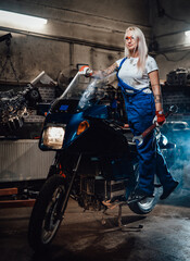 Beautiful female mechanic with tattooed hands wearing work overalls posing on her sportbike in garage or workshop